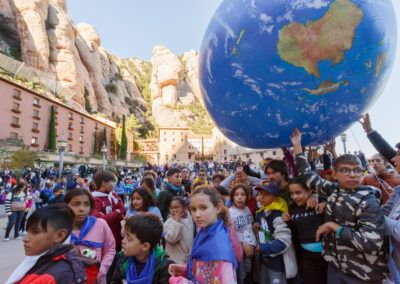Niños y niñas participando en un evento cultural al aire libre, con una gran esfera del planeta Tierra, en el entorno de Montserrat.