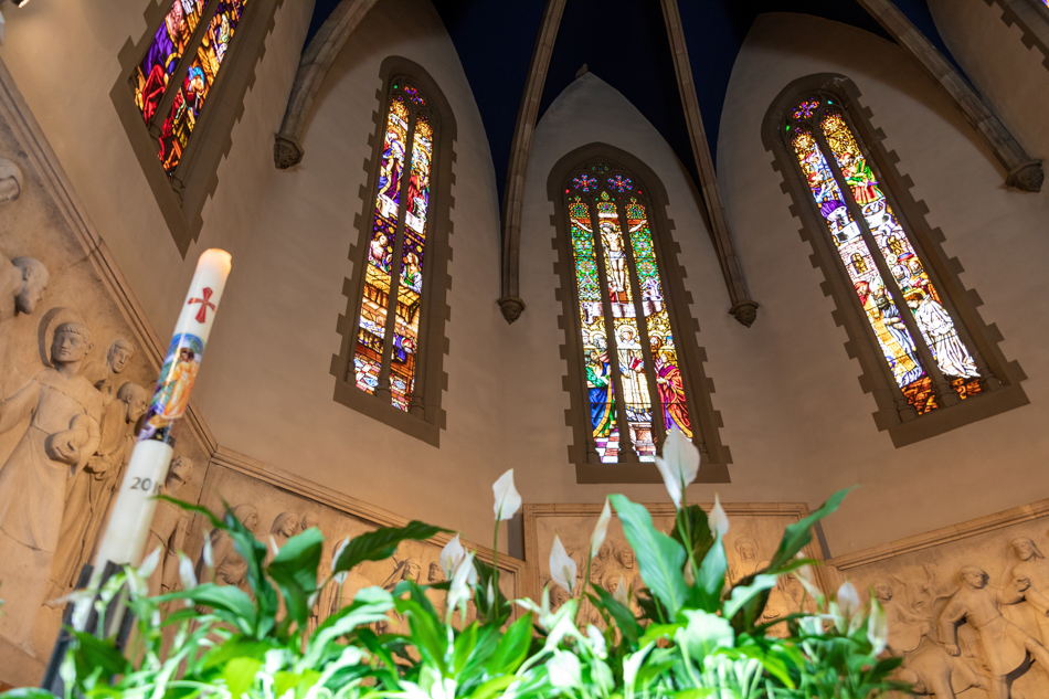 Interior de una iglesia con vidrieras, cirio y decoración floral durante un reportaje de fotografía de bautizo en Granollers, Vallès Oriental, por Elenirc Fotografia.