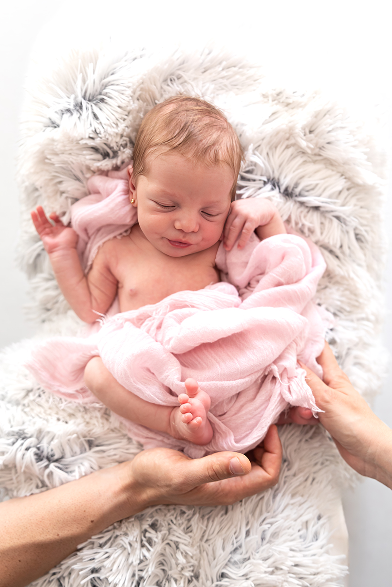 Bebé recién nacido envuelto en una muselina rosa y sostenido por unas manos durante una sesión de fotografía newborn en estudio en Mollet del Vallès, por Elenirc Fotografia.