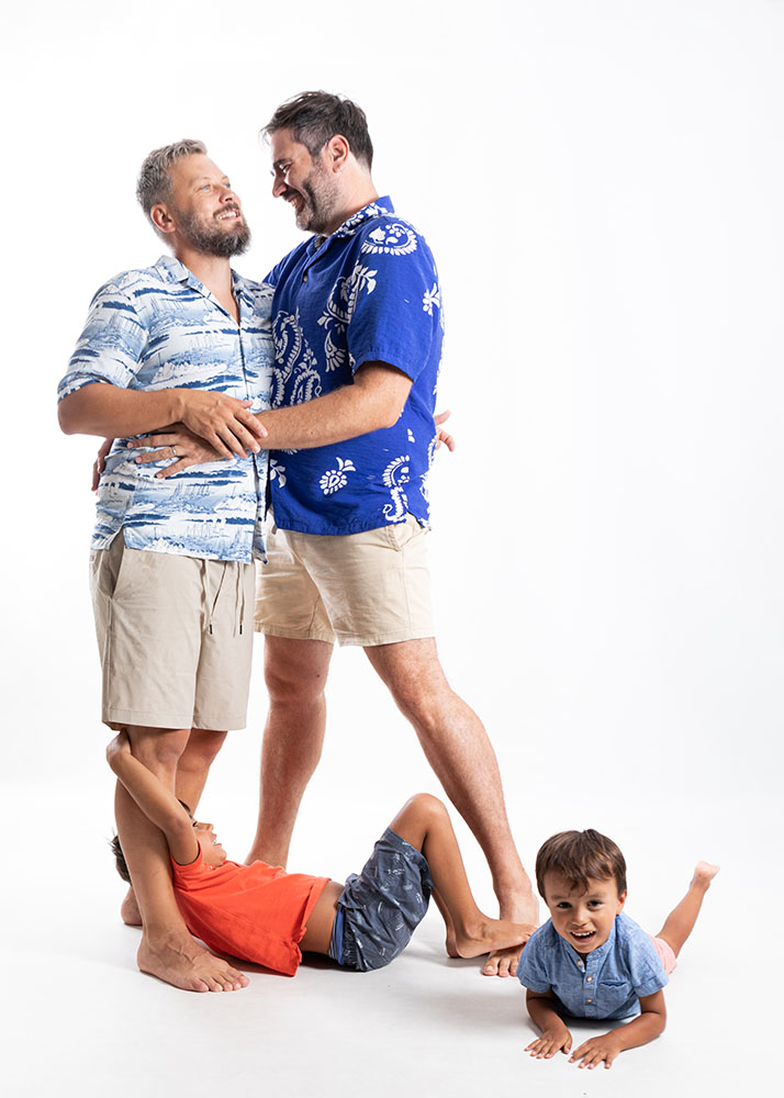Sesión de fotografía familiar con dos padres y sus hijos. Papá y papi abrazados en el estudio mientras sus dos hijos pequeños juegan