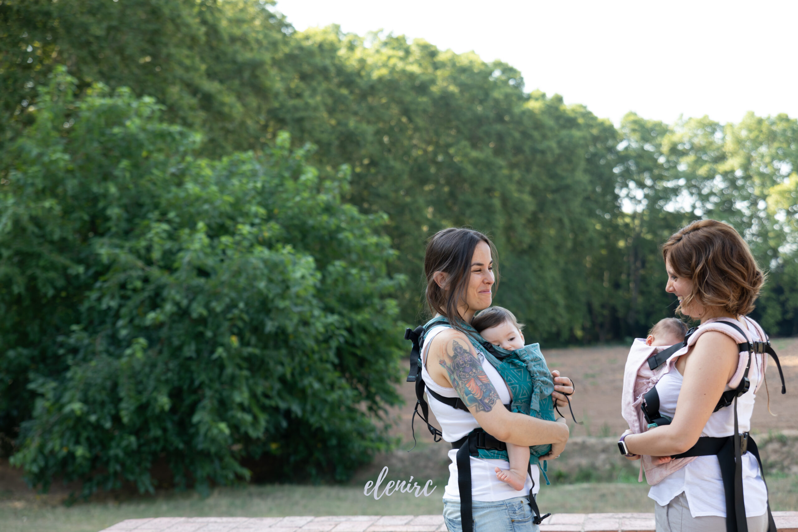 Maternidad y porteo de bebés al aire libre · elenirc Dos mujeres conversan mientras llevan a sus bebés en portabebés ergonómicos durante un paseo al aire libre.