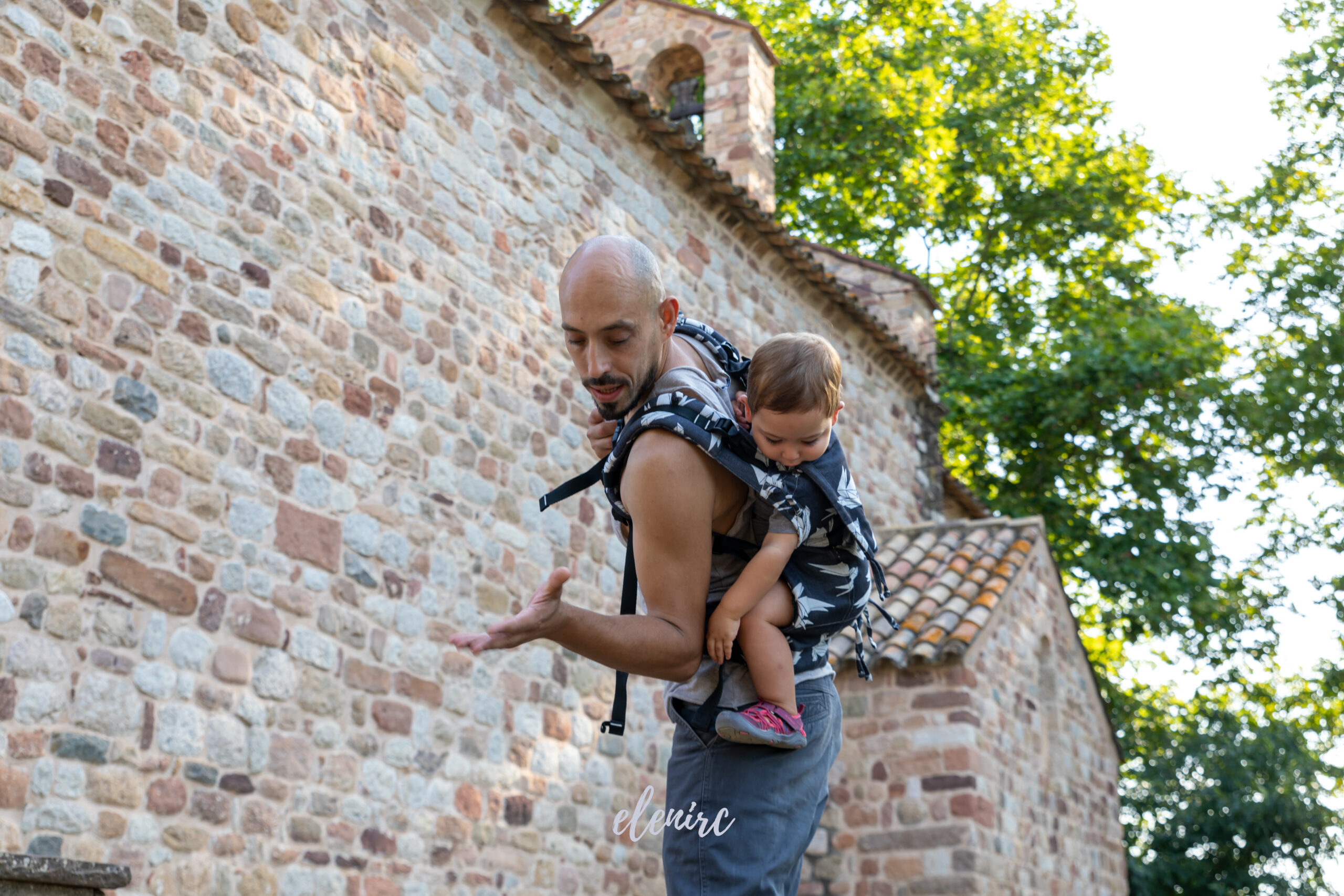 Porteo a la espalda con mochila portabebés · elenirc Padre caminando junto a un muro de piedra con su bebé a la espalda en una mochila portabebés. Fotografía documental de Elena Rubio · elenirc.