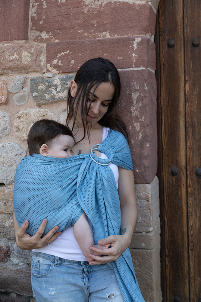 Porteo con fular azul · Elena Rubio · elenirc Madre mirando a su bebé mientras lo lleva en un fular azul, junto a una puerta de madera y un muro de piedra.