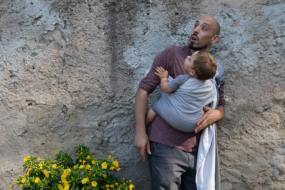 Paternidad y porteo · Reportaje para tienda de maternidad Padre con su bebé en un fular gris junto a un muro de piedra. Fotografía de porteo para un proyecto comercial de maternidad y crianza.
