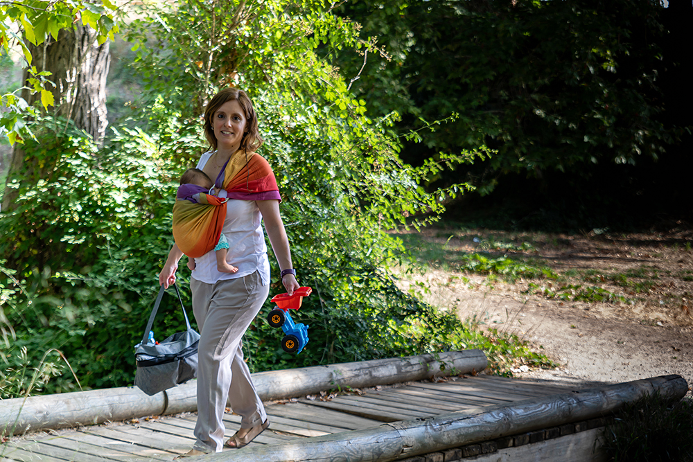 Fotografía de porteo para marcas de maternidad · elenirc Madre caminando por una pasarela de madera con su bebé en un fular de porteo de colores. Reportaje fotográfico de Elena Rubio.