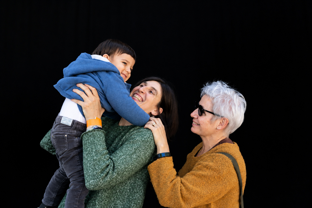 Dos mamás abrazan a su bebé sonriente, que extiende los brazos hacia la cámara. Retrato de la colección Som famílies a tot arreu, de Elena Rubio · elenirc.