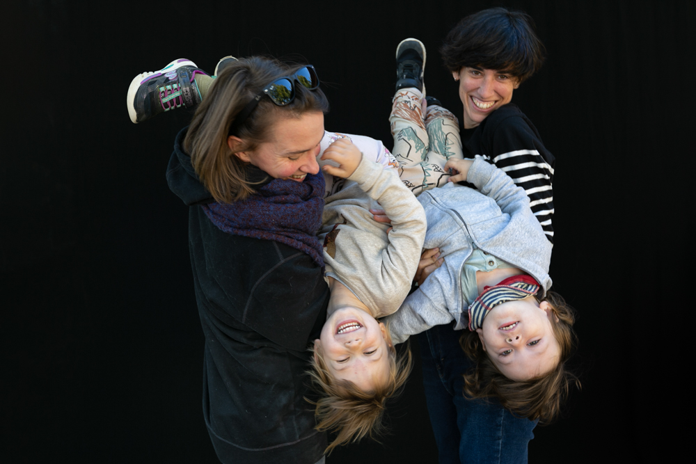 Dos mamás jugando con sus dos hijos, que ríen boca abajo mientras los sostienen en brazos. Una colección de abrazos de familias LGTBI+ durante la Castañada de FLG. Fotografía de Elena Rubio · elenirc.