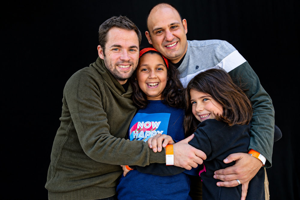 Dos papás se abrazan con sus dos hijas durante la celebración de la Castañada de FLG · Famílies LGTBI+. Retrato de familia homoparental de Elena Rubio · elenirc.