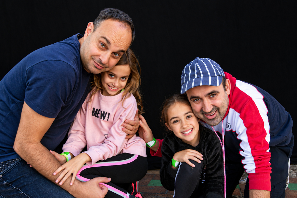 Dos papás abrazan a sus dos hijas frente a un fondo negro. Retrato de familia homoparental durante la Castañada de FLG. Fotografía de Elena Rubio · elenirc.
