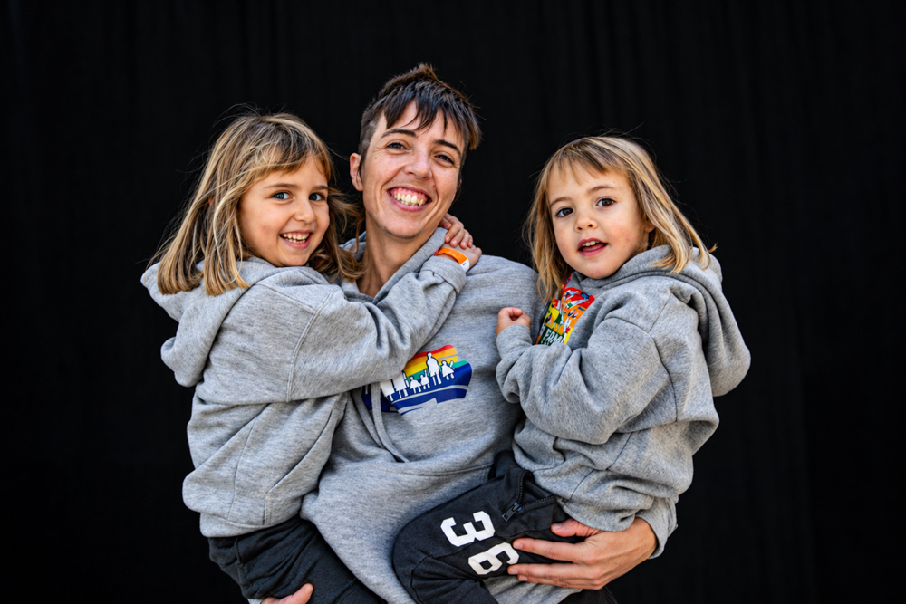 Una mamá recibe el abrazo de sus dos hijas, que sonríen ante la cámara. Retrato familiar de la colección Som famílies a tot arreu, fotografiado por Elena Rubio · elenirc.