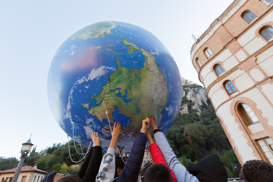 Manos de varios participantes sostienen un gran globo terráqueo en una actividad colectiva al aire libre. Fotografía de Elenirc Fotografia.