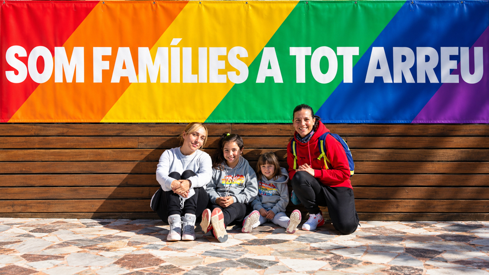 Dos mamás con sus dos hijas ante una pancarta arcoíris con el lema Som famílies a tot arreu. Retrato de familia homoparental durante la celebración de FLG, por Elena Rubio · elenirc.
