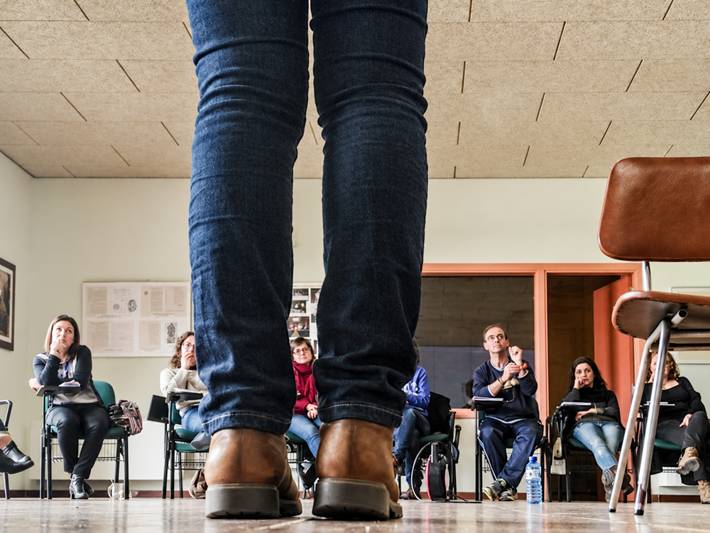 Perspectivas de un taller · Fotografía documental Elena Rubio elenirc Plano bajo de las piernas y zapatos de una persona, con los asistentes a un taller de gestión emocional al fondo. Fotografía documental de marca personal Elena Rubio · Elenirc Fotografia.